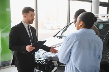 Araba satıcısı, Afrika kökenli Amerikalı çifte yeni araba özelliklerini sunuyor. Modern galeri galerisinde alışverişi tartışıyorlar.