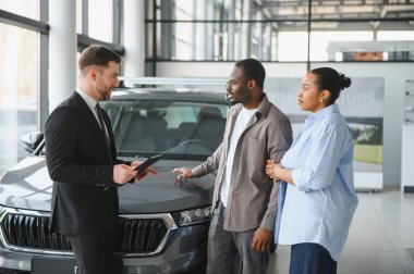 Afrika kökenli Amerikalı çifte yeni otomobil sunan profesyonel satıcı, modern araba galerisinde alışverişi tartışıyor.