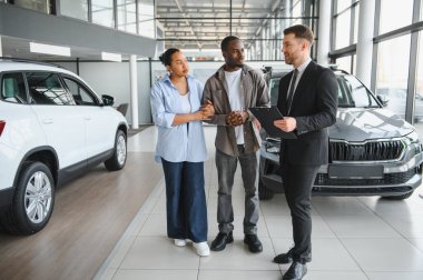 Araba satıcısı elinde not panosuyla, modern araba galerisindeki Afrikalı Amerikalı çifte yeni bir araç sunuyor.