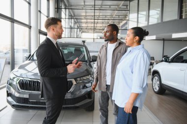 Araba satıcısı tablet kullanıyor ve araba galerisindeki bir çifte yeni bir araba gösteriyor. Özellikleri ve özellikleri tartışıyorlar.