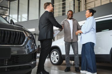 Araba satıcısı mutlu bir çiftle el sıkışıyor, modern bir galeride yeni bir araba alımı yapıyor.