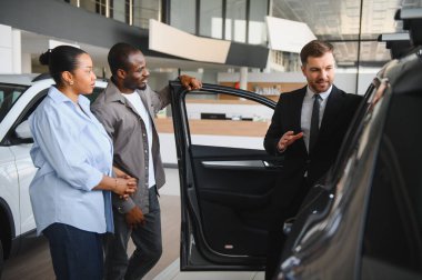 Araba satıcısı, modern bir araba galerisinde Afro-Amerikan bir çifte yeni bir arabanın özelliklerini sunuyor.