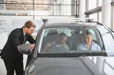 Araba satıcısı içeride oturan Afrikalı Amerikalı bir çifte yeni bir aracın özelliklerini gösteriyor. Modern bir araba galerisinden bir şey almayı düşünüyorlar.