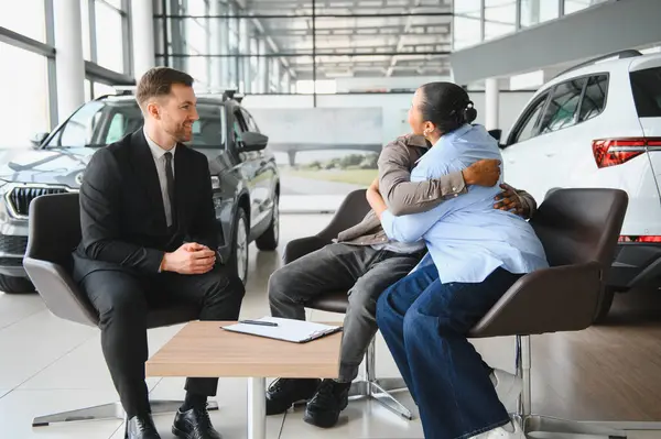 Heyecanlı çift kucaklaşıyor, yeni araba alımlarını modern bir galeride bir araba satıcısıyla kutluyorlar.