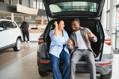 Yeni arabalarının bagajında oturan mutlu çift, bir araba galerisinde alışverişlerini kutluyorlar.