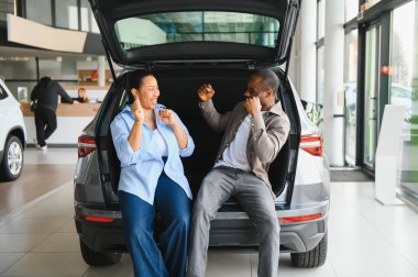 Yeni arabalarının bagajında oturan heyecanlı çift, alışverişlerini neşeli ifadelerle kutluyorlar.