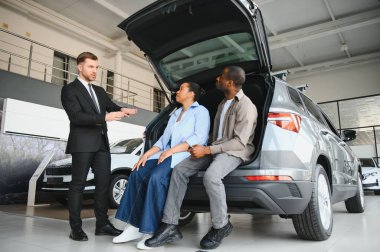 Araba satıcısı, bir galeride açık bagajda oturan bir çifte yeni arabanın özelliklerini sunuyor.