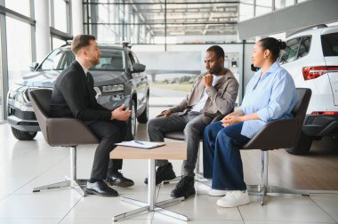 Araba satıcısı oturuyor ve bir galeriden yeni bir araba almayı düşünen Afrikalı Amerikalı bir çiftle sohbet ediyor.