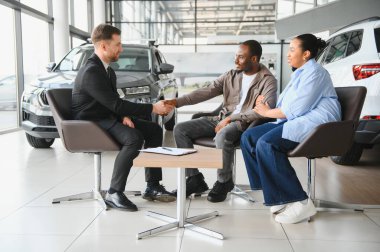 Satıcı, Afro-Amerikan çiftiyle el sıkışıyor. Modern bir bayiliğin içinde yeni bir araba için anlaşma yapıyor.