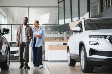 Gülümseyen bir çift, parlak bir araba galerisinde yürürken seçenekleri ve özellikleri tartışırken mutlu bir şekilde yeni otomobillerini seçiyorlar.