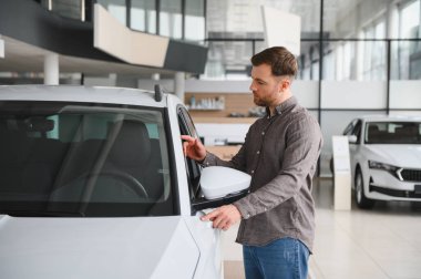 Otomobil galerisinde yeni bir beyaz arabayı inceleyen, araba gövdesine ve yan aynasına dokunan, satın alma seçeneklerini göz önüne alan bir adam.