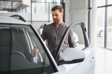 Adam, lüks seçeneklerle dolu modern bir galeride yeni bir araç seçerken arabanın kapısını açıyor ve içini inceliyor.