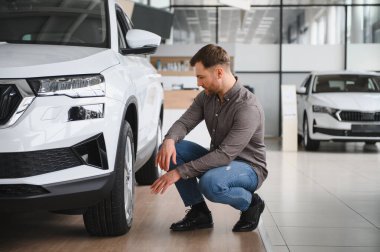 Araba galerisinde diz çöküp araba lastiğini yakından inceleyen bir adam yeni bir araç almayı dikkatlice düşünüyor.
