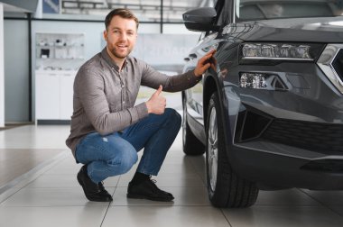 Bir araba galerisinde yeni gri bir cipin yanında çömelmiş cesede dokunup baş parmak hareketi yapan mutlu bir adam.