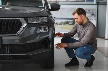 Adam çömelmiş, araba galerisinde yeni gri bir SUV 'u inceliyor.