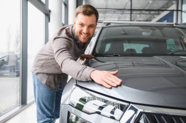 Heyecan ve olasılıklarla dolu canlı bir galeride yeni bir arabanın kaputuna dokunmanın tadını çıkaran mutlu bir adam.