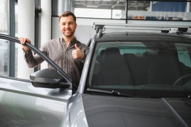 Bir galeride yeni bir arabanın kapısını açarken gülümseyen adam, satın almasından duyduğu memnuniyeti dile getiriyor.