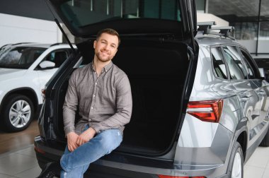 Yeni arabanın açık bagajında oturan mutlu adam araba galerisindeki yerleri ve özellikleri kontrol ediyor.