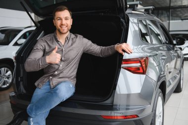 Açık araba bagajında otururken mutlu müşteri el kol hareketi yapıyor, galeride yeni bir otomobil seçiyor, satın almaktan memnun.