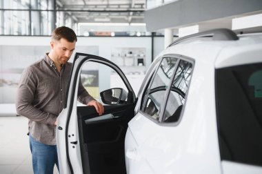 Bir galerinin içinde beyaz bir arabanın kapısını açan adam, satın almadan önce iç mekanı inceliyor.