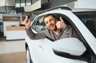 Araba anahtarlarını ve baş parmağını gösteren heyecanlı bir adam galeride yeni araba alımını kutluyor.