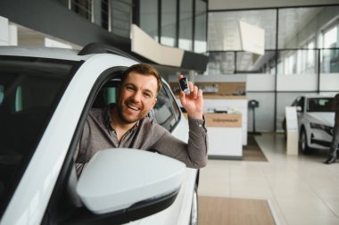 Gülümseyen adam, galeride yeni alınmış arabasında otururken yeni araba anahtarlarını gösteriyor.