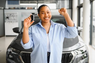 Yeni araba alımını kutlayan mutlu bir müşteri modern, şık bir galeride, parlak ışıklarla dolu bir arabanın anahtarlarını tutuyor.