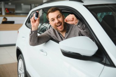 Bir araba galerisinde yeni arabasında oturan mutlu adam araba anahtarlarını gururla sergiliyor ve başarılı alımını kutluyor.