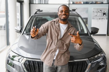 Gururlu bir Afrikalı Amerikalı araba galerisinde yeni arabasının önünde dikilirken araba anahtarlarını gösteriyor.