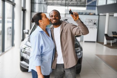 Afro-Amerikalı çift öpüşüyor ve yeni araba anahtarlarını gösteriyor, araba galerisinde alışverişlerini kutluyorlar.