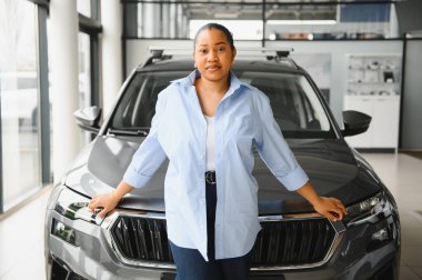 Kendine güvenen Afrikalı Amerikalı kadın galeride yeni bir araba seçiyor, gri bir cipin kaputuna yaslanıyor.