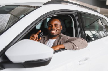 Gülümseyen adam, yeni bir araba galerisinde otururken arabanın anahtarlarını tutuyor. Son satın alımının heyecanıyla eğleniyor.