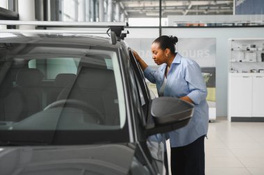 Afro-Amerikalı bir kadın satış mağazasında yeni bir arabayı teftiş ediyor, satın alma seçeneklerini göz önünde bulundurarak.