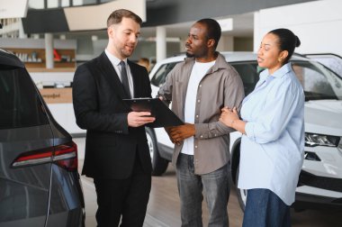 Araba satıcısı elinde bir pano tutarken canlı bir araba galerisinde Afro-Amerikan bir çifte araçları gösteriyor ve seçenekleri tartışıyor.