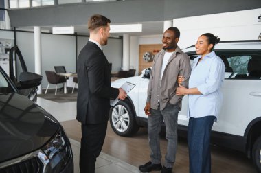 Araba satıcısı elinde not panosuyla gülümseyen Afro-Amerikan çifte yeni bir araba gösteriyor.