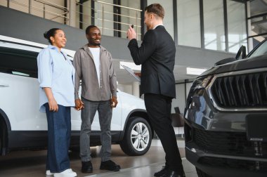 Araba satıcısı, modern bir bayiliğin içinde Afrikalı Amerikalı bir çifte yeni bir araba sunuyor.