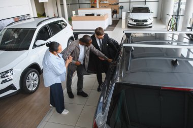 Satıcı, bir araba galerisinde Afro-Amerikan bir çifte doğru aracı seçmede yardım ve rehberlik eden yeni bir arabanın özelliklerini gösteriyor.