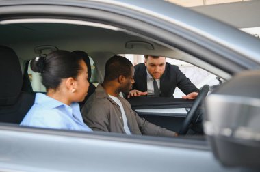 Araba satıcısı, yeni bir aracın özelliklerini galeriden araba almak isteyen Afrikalı Amerikalı bir çifte gösteriyor.