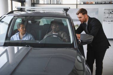 Araba satıcısı içeride oturan Afro-Amerikan bir çifte yeni bir arabanın özelliklerini gösteriyor. Modern bir galeride alım opsiyonlarını tartışıyorlar.