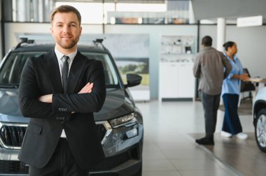 Müşteriler galeride otomobili seçerken, kendinden emin bir satıcı arabanın önünde çapraz kollarla poz veriyor.