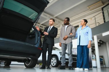 Araba satıcısı, yeni bir arabanın bagajını Afro-Amerikan bir çifte tanıtıyor. Kalabalık bir araba galerisinde.