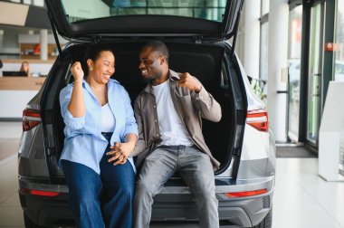 Bir galeride arabanın bagajında oturmuş gülümseyen bir çift yeni arabalarını seçerken bir satıcıyla kapışıyor.