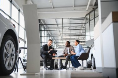 Gülümseyen araba satıcısı, modern bir araba galerisindeki masada oturan genç bir Afrikalı Amerikalı çifte kontrat gösteriyor.