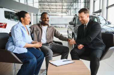 Gülümseyen çift bir satıcıyla el sıkışıyor. Parlak ve hoş bir galeride yeni bir araba satın alınmasını kutluyorlar.