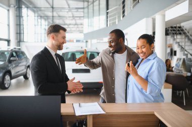 Heyecanlı çift bir araba satıcısıyla el sıkışıyor galeride başarılı bir şekilde yeni bir araç satın alınmasını kutluyorlar.