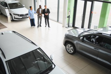 Araba satıcısı, parlak ve ferah bir araba galerisinde yeni araçlar sunarken Afro-Amerikan bir çiftle takılıyor.