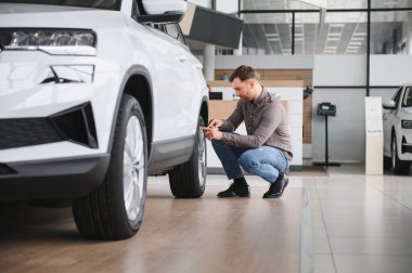 Bir araba galerisinde çömelmiş ve yeni bir SUV 'un lastiklerini inceliyor.