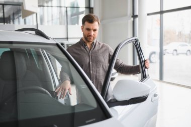 Yakışıklı bir adam galeride yeni bir arabayı inceliyor, kapıyı açıyor ve direksiyona dokunuyor.