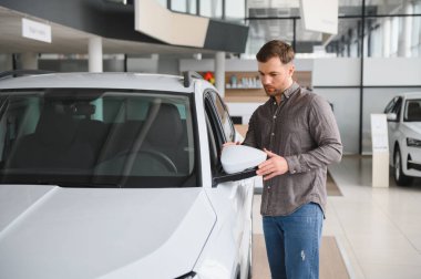 Araba galerisindeki beyaz bir arabanın yan aynasını inceleyen bir adam.