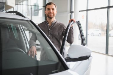 Mutlu adam yeni bir araba seçiyor, kapıyı açıyor ve modern bir galeride test sürüşüne hazırlanıyor.